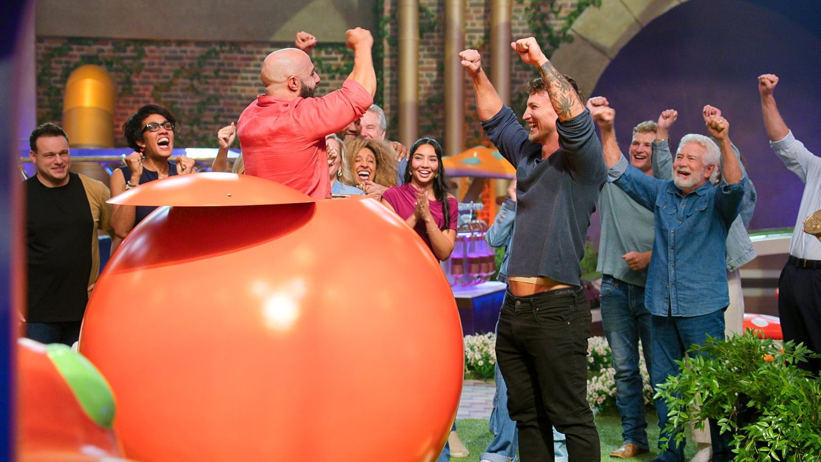 The cast cheering with fists raised beside a giant orange sphere, Nick Spinelli smiling on the far left