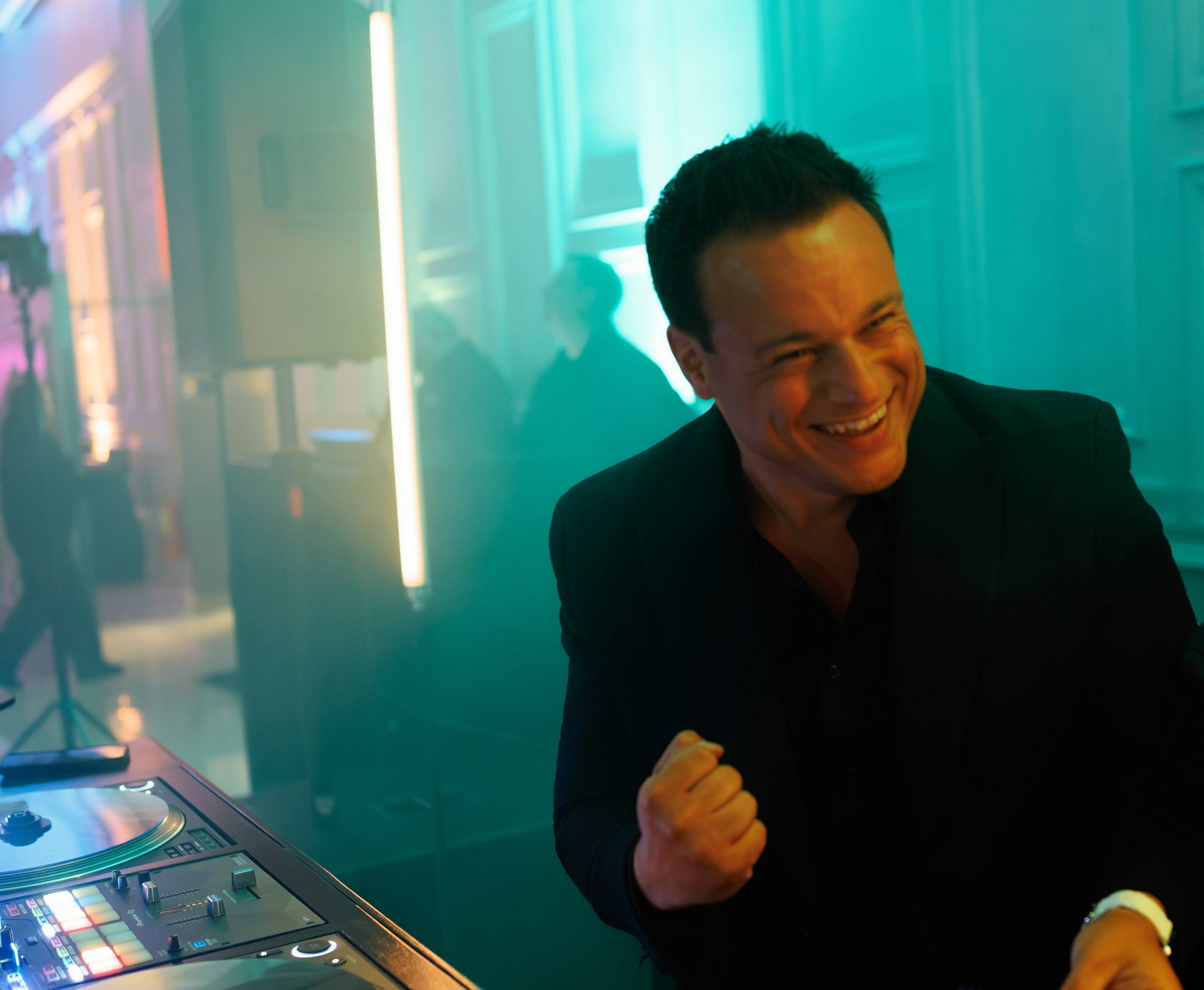 Nick Spinelli laughing and pumping his fist behind the decks under teal uplighting